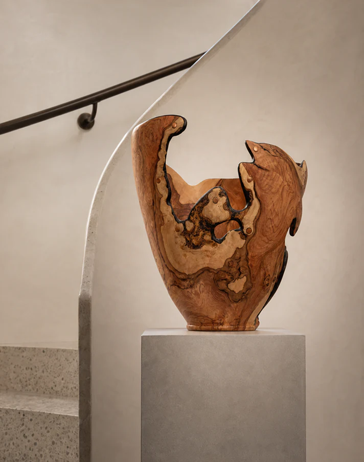 Tall sculptural Wild Olive vessel displayed on a stone plinth beside a curved staircase.